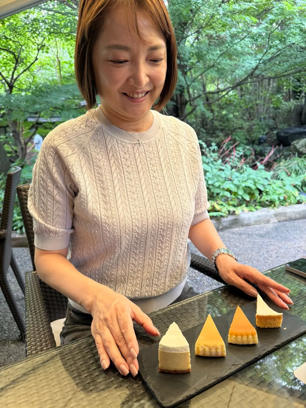 笑顔の女性が、黒い石板の上に並んだ４つのチーズケーキを眺めている。女性はベージュのケーブル編みトップスを着ており、左手首に腕時計をしている。背景には緑豊かな庭園が見える。