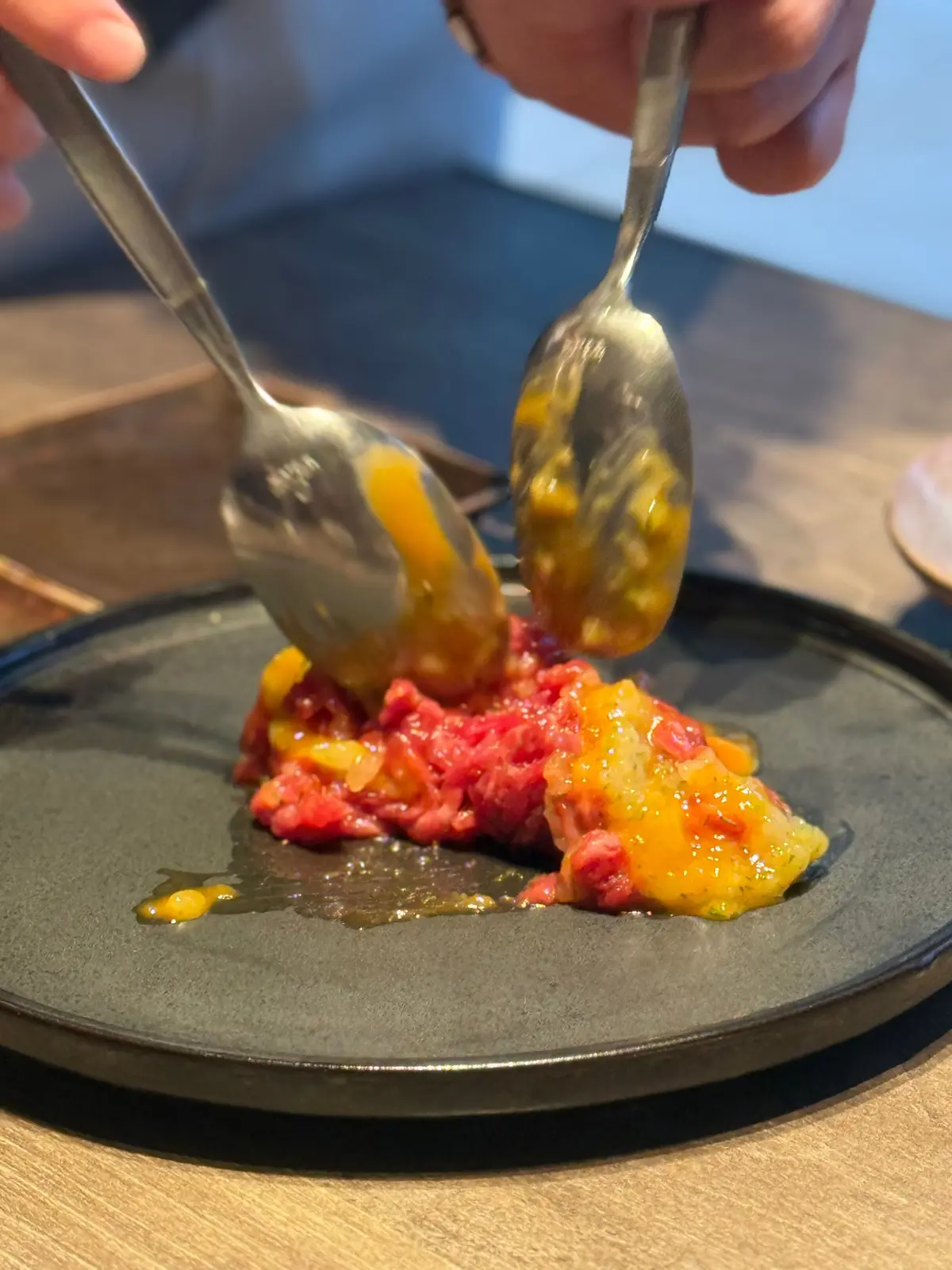 黒い皿に盛り付けられた、赤身の肉を細かく刻んだ料理。2つの銀色のスプーンで、黄色いソースがかけられている。