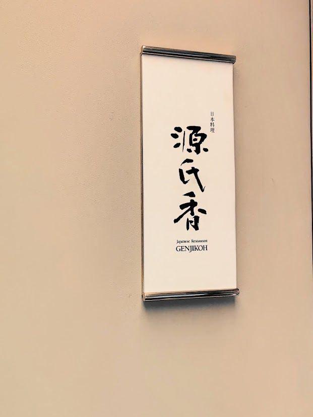 淡いベージュの壁に、銀色のフレームで囲まれた縦長の看板。中央に「源氏庵」と書かれ、下部に「Japanese Restaurant GENJIKO」と英語表記がある。