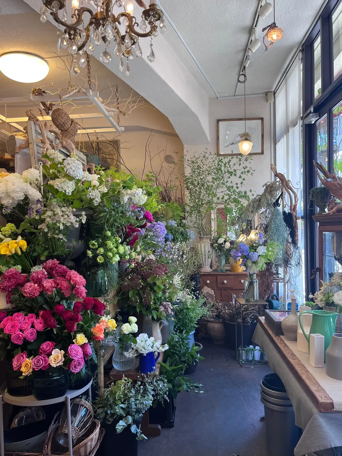 花屋の店内。多様な花が色とりどりに並び、窓から自然光が差し込む。天井にはシャンデリアが輝き、さまざまな花瓶や装飾品が置かれている。