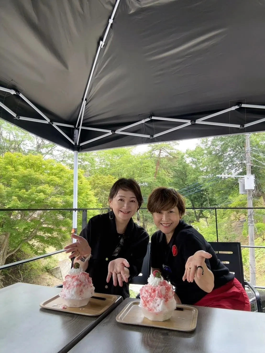 2人の女性が、木製トレイにのったイチゴのシャーベットを手に、屋外のテント下で笑顔でポーズしています。背景には緑豊かな木々が広がっています。