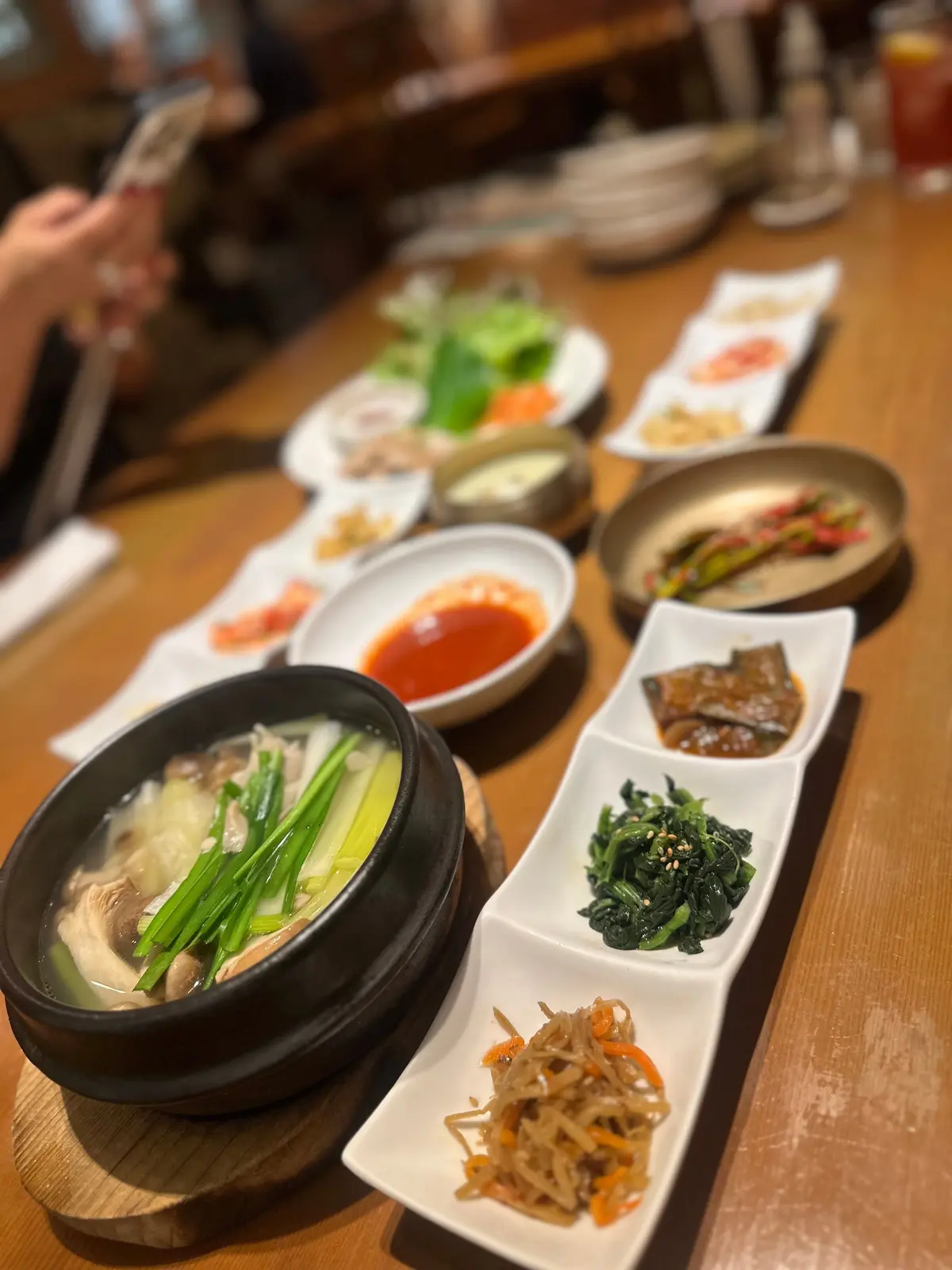 木製のテーブルに並べられた韓国料理の盛り合わせ。石製の鍋にはきのこや野菜が煮込まれ、白磁の小皿には、ナムルやキムチ、赤色のソースなどが彩り豊かに盛り付けられている。