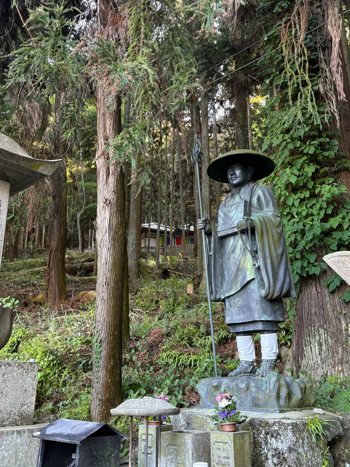 青銅の僧侶像が森の中の石の上に立ち、笠と杖を持っている。周囲には石の灯篭や花が置かれている。