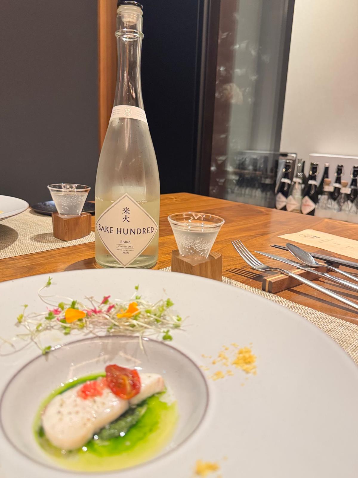木製のテーブルに置かれた白い皿に、魚料理と彩り豊かな野菜が盛り付けられている。近くには「Sake Hundred」と書かれた日本酒のボトルとグラスが置かれ、フォークとナイフが並んでいる。背景には暗い壁と、ぼやけたテーブル席が見える。
