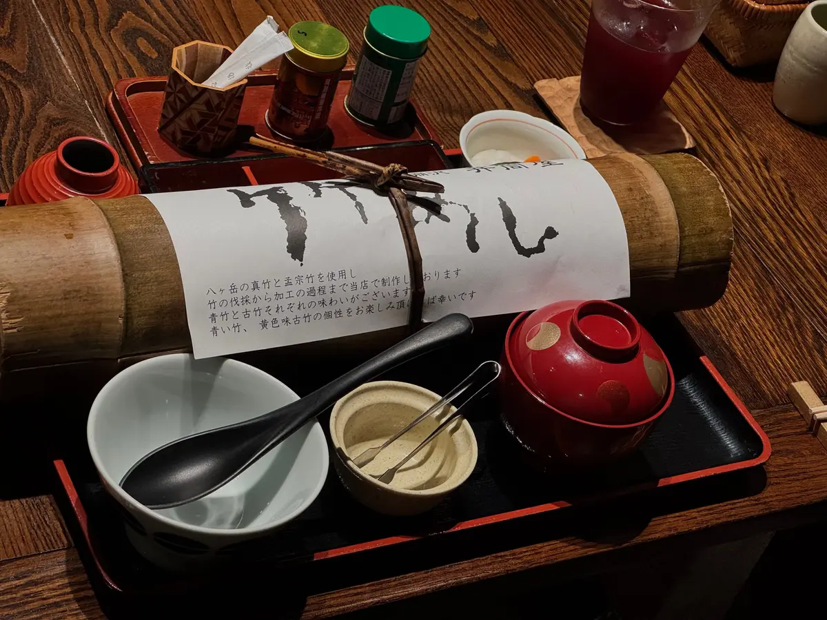 木製のトレイに、ラーメンの器、スプーン、醤油差し、ラー油などが置かれている様子。中央には、手書き文字の書かれた巻物がある。背景には、茶色い食器が並んでいる。