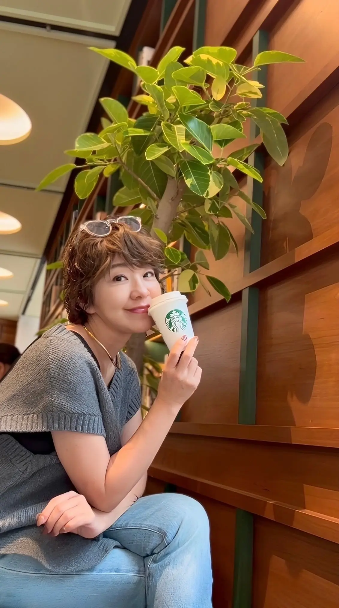 カフェでスターバックスのカップを持つ女性。茶色の短髪で、グレーのトップスとジーンズを着用。背景には木製の階段と観葉植物が見える。