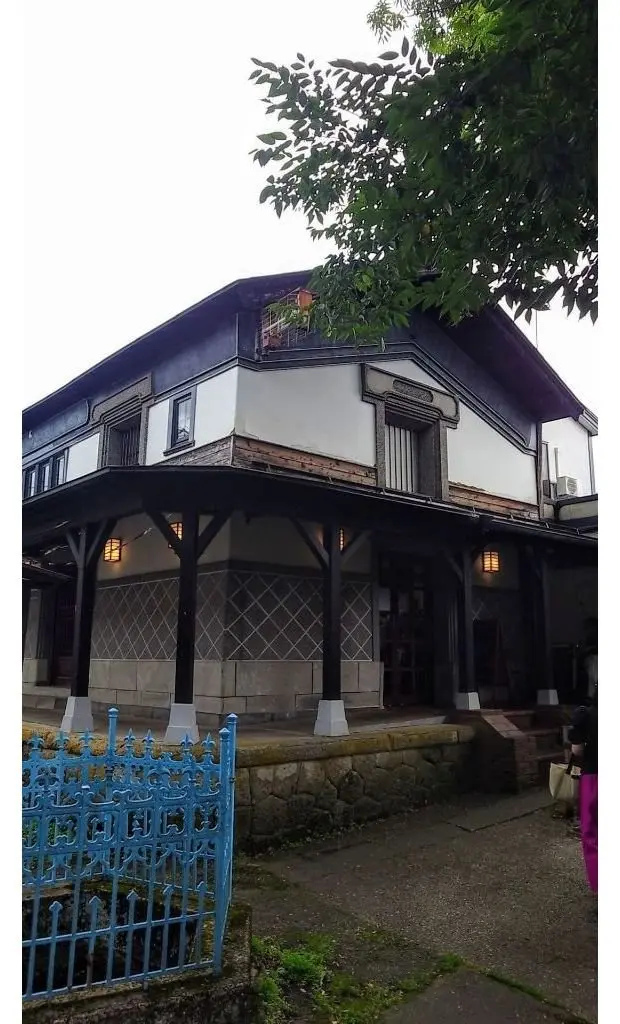 木造の2階建て建築。白と黒のコントラストが際立つ外観で、庇と柱が特徴的。手前に青い鉄柵があり、緑の植物が生い茂っている。