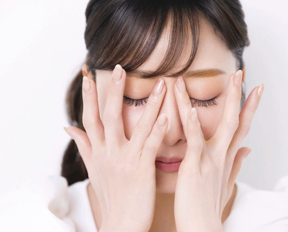 白シャツを着た女性が目を閉じ、両手で顔を覆っている。茶色の前髪が額にかかり、背景は白。