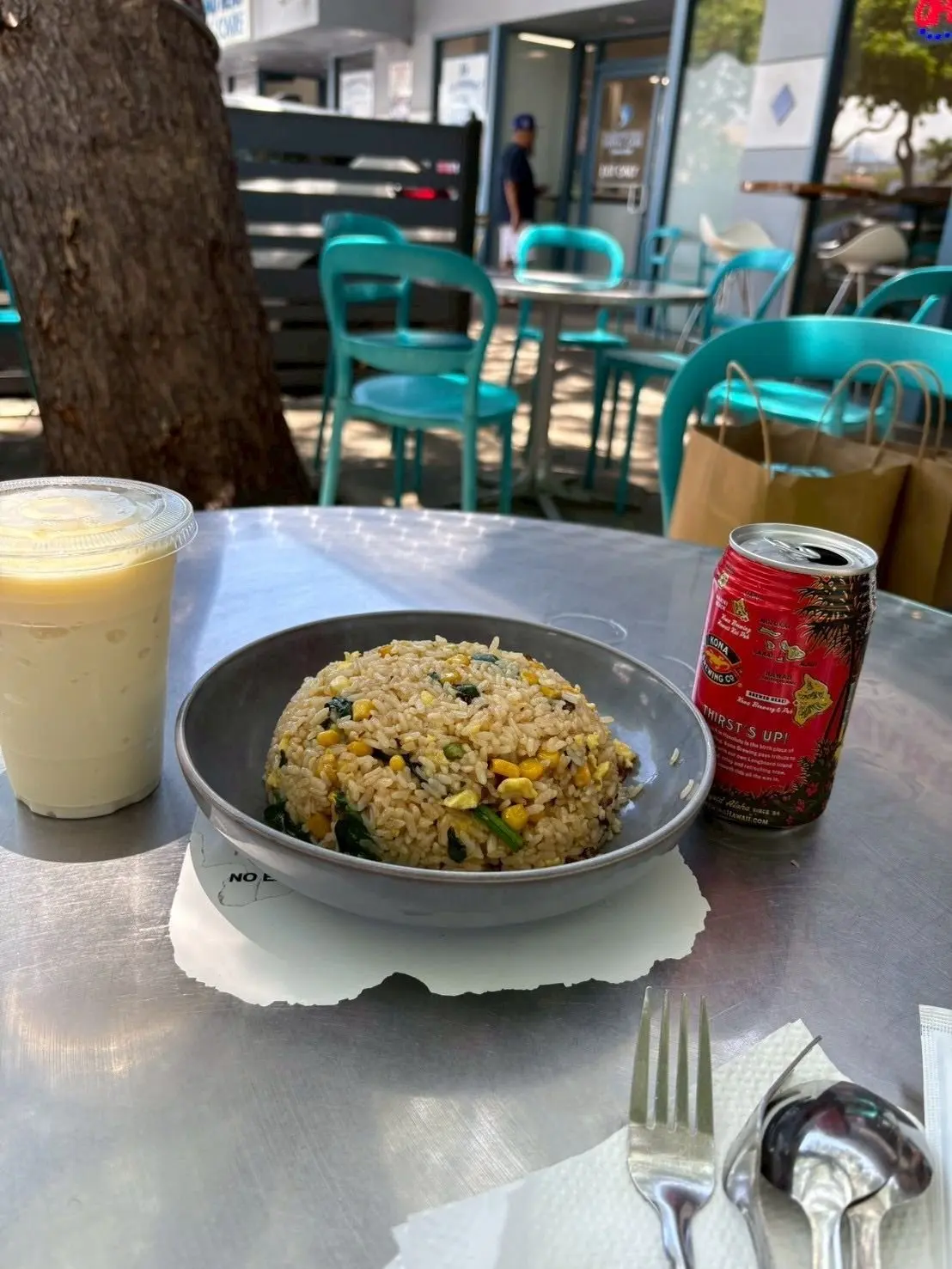 テーブルに置かれた、タイ料理のチャーハンとアイスティー、コナ Brewing Co.の缶ビール。青い椅子が並ぶ屋外のカフェ。