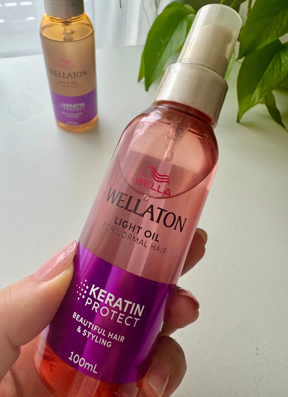 ウェラ Wellaton ライトオイル、100mlのボトル。紫色のラベルに「KERATIN PROTECT」の文字。背景には緑の葉と、同じブランドのオイルボトルが写っている。