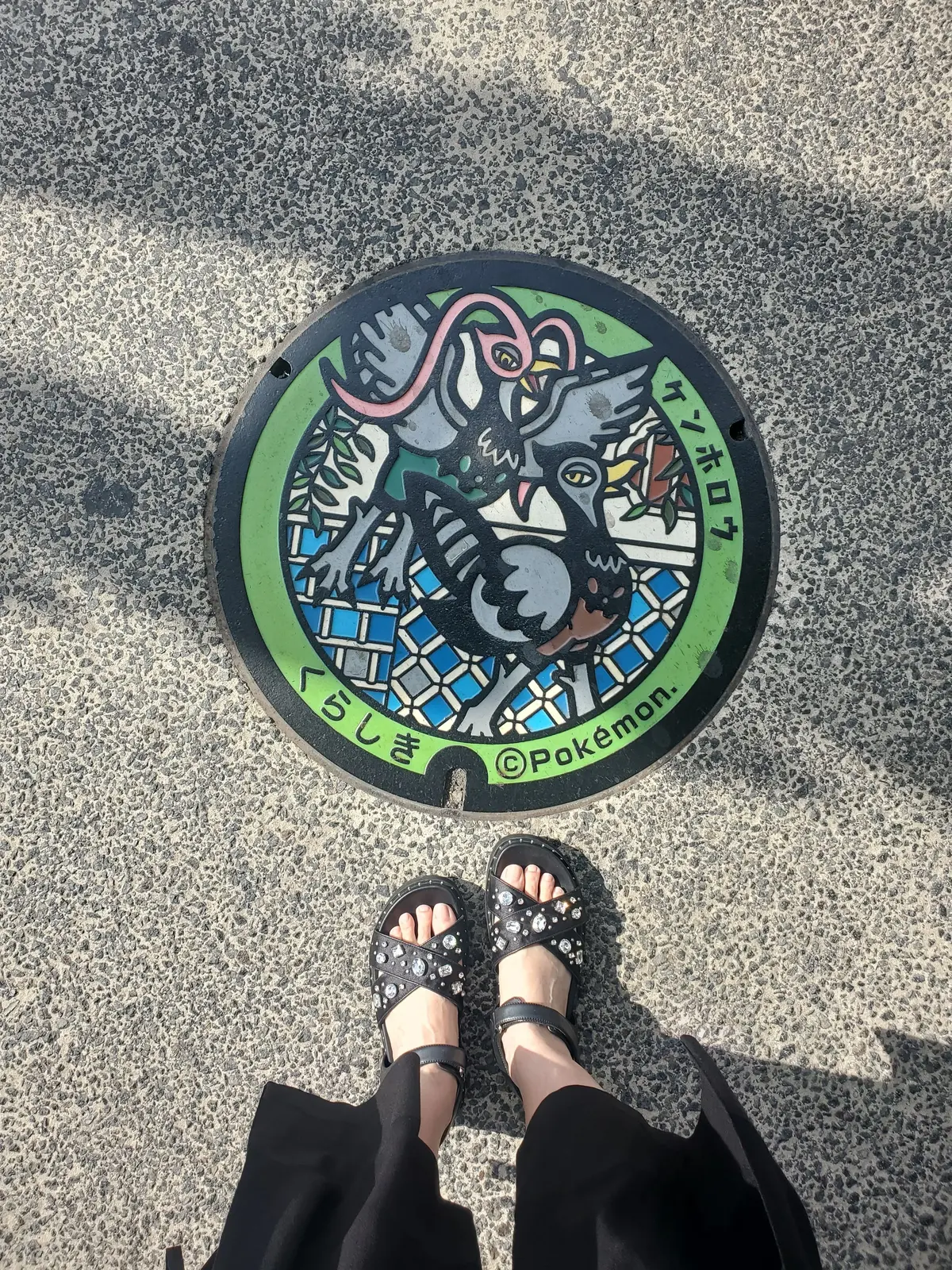 道路上のマンホールに描かれたポケモン「ケンタロス」のデザイン。緑と茶色の縁取りで、中央には青いケンタロスが描かれ、周囲に幾何学模様が配置されている。©Pokémonのロゴマークがある。