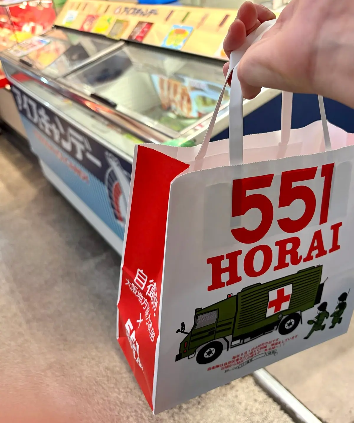 白地に赤字で「551 HORAI」と記載された紙袋。中央には緑色のトラックのイラストが描かれている。背景には食品を陳列した冷蔵ケースが見える。