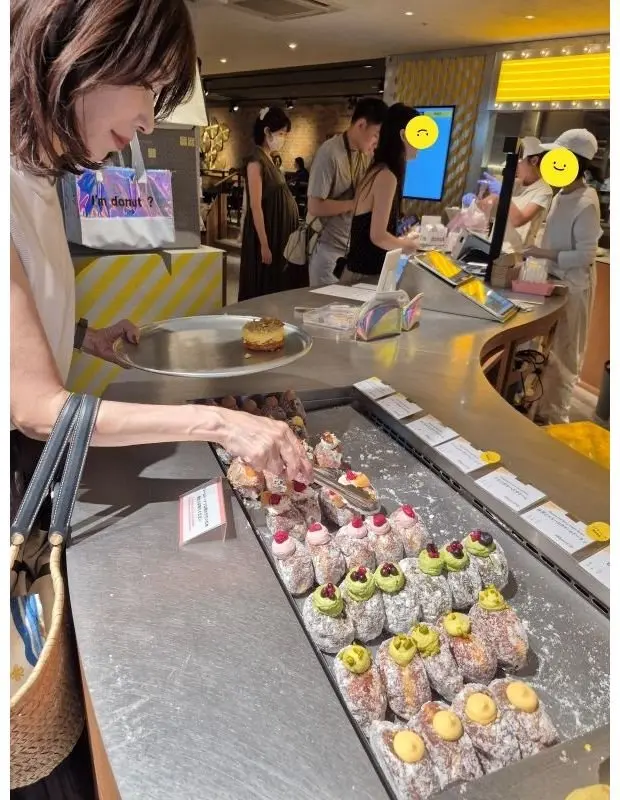 ドーナツ店で女性がドーナツを選んでいる様子。ステンレス製のカウンターに、様々な種類のドーナツが並び、女性はトングでドーナツを手に取ろうとしている。背景には、他の客や店員が見える。