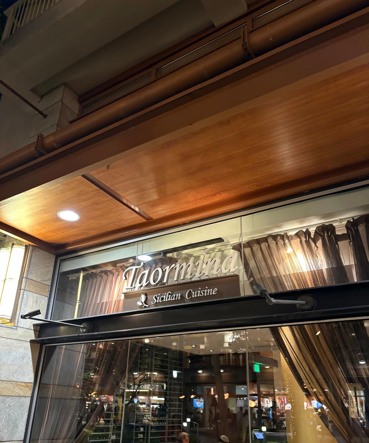 Taorminaと書かれた看板が、木目調の天井の下に設置されたレストランの入り口。ガラス張りの窓から内部の様子がうかがえる。