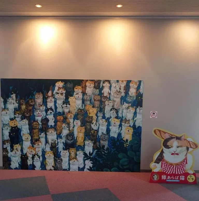 猫がびっしりと描かれた大きな絵画。様々な毛色と表情の猫が密集し、手前に緑の葉が見える。右下には猫のイラストと「磯あらし猫」の文字が入った赤い看板。