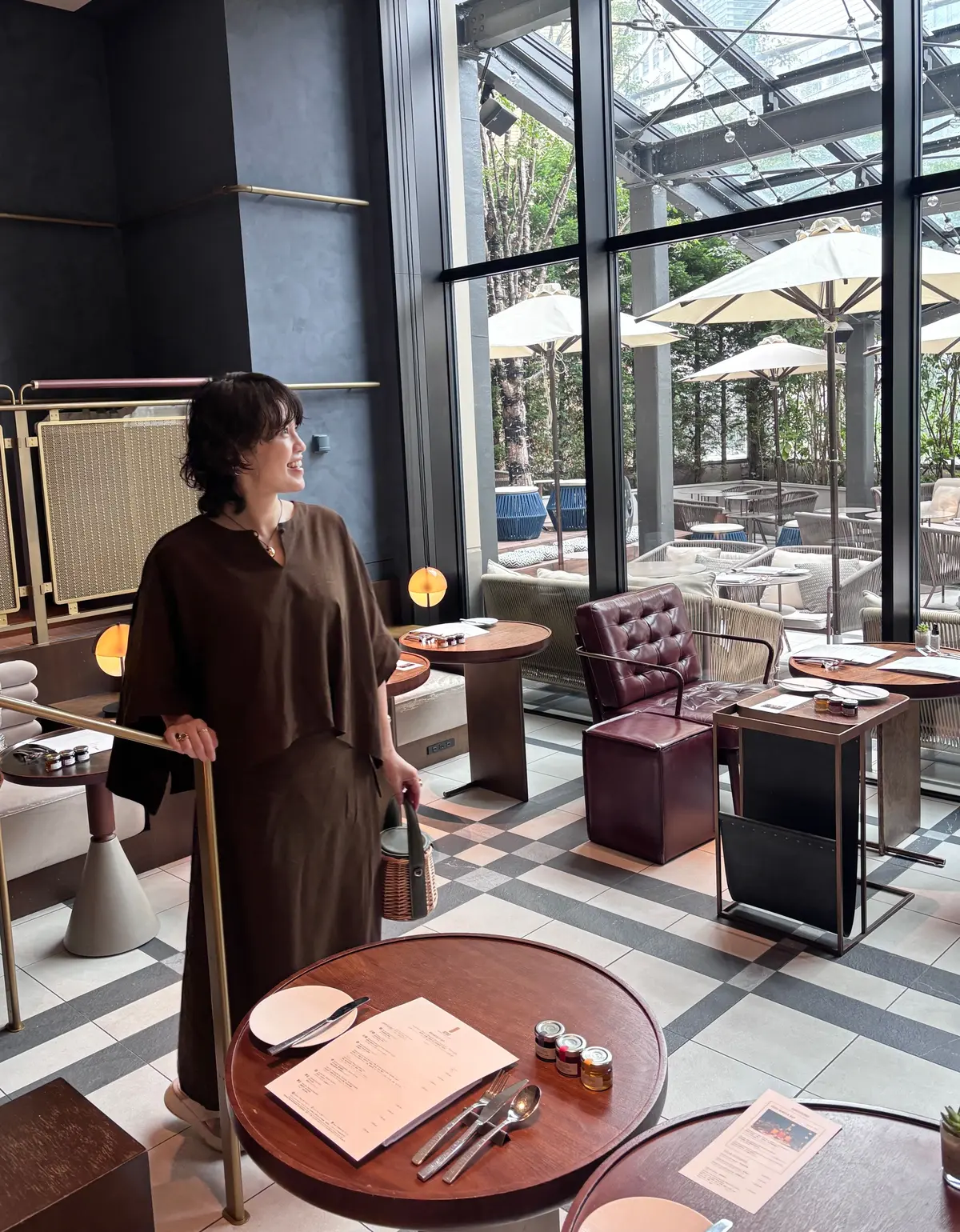 茶色のワンピースを着た女性が、レストランのテーブルの傍に立っている。背景には、大きな窓から緑豊かな屋外のテラス席が見える。テーブルにはメニューやカトラリーが置かれ、床は白黒のタイル張り。