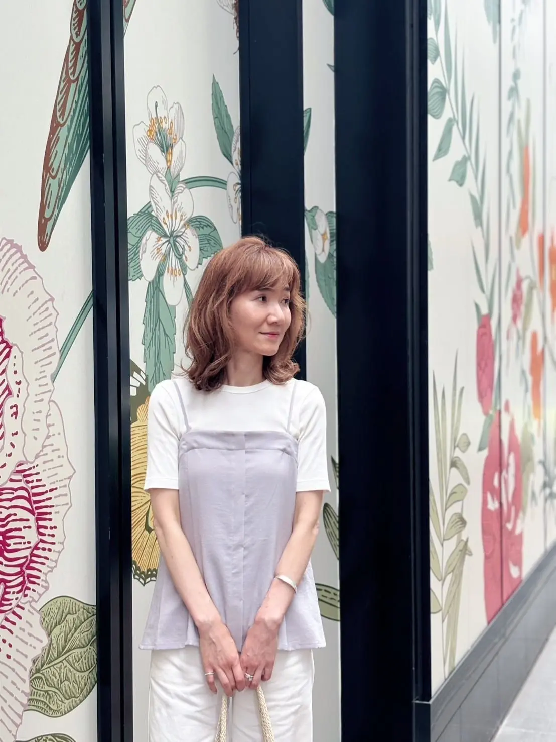 花の模様が描かれた壁の前で、白いトップスとグレーのサロペットを着た女性が立っている