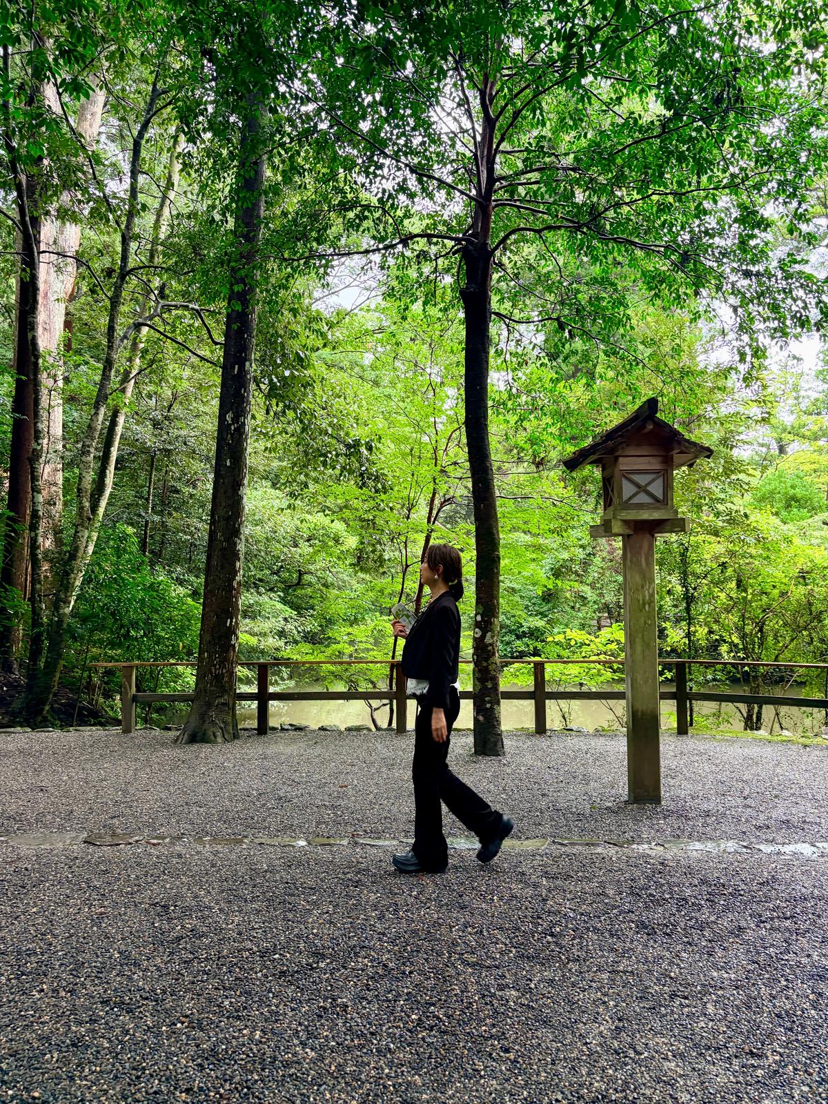 石畳の道に、黒い服を着た女性が杖をついて歩いている。背景には緑豊かな木々と、木製の灯籠が見える。