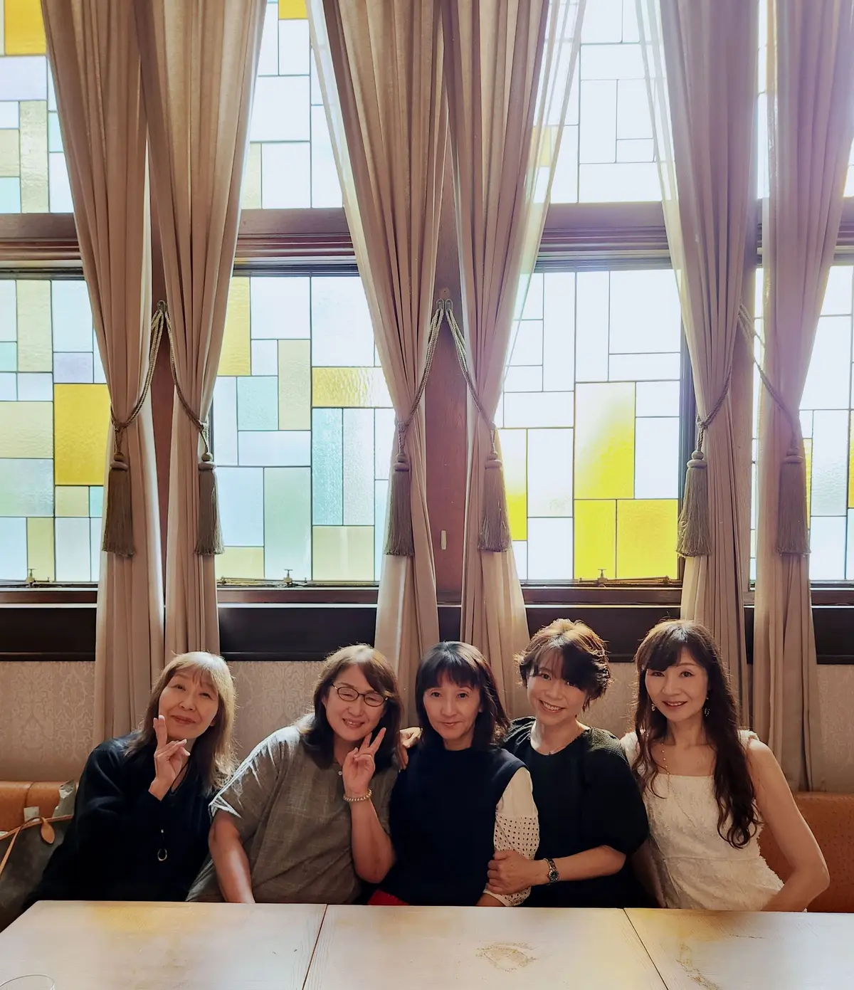 5人の女性が木製のテーブルに座り、窓のステンドグラスとベージュのカーテンの背景に笑顔で並んでいる