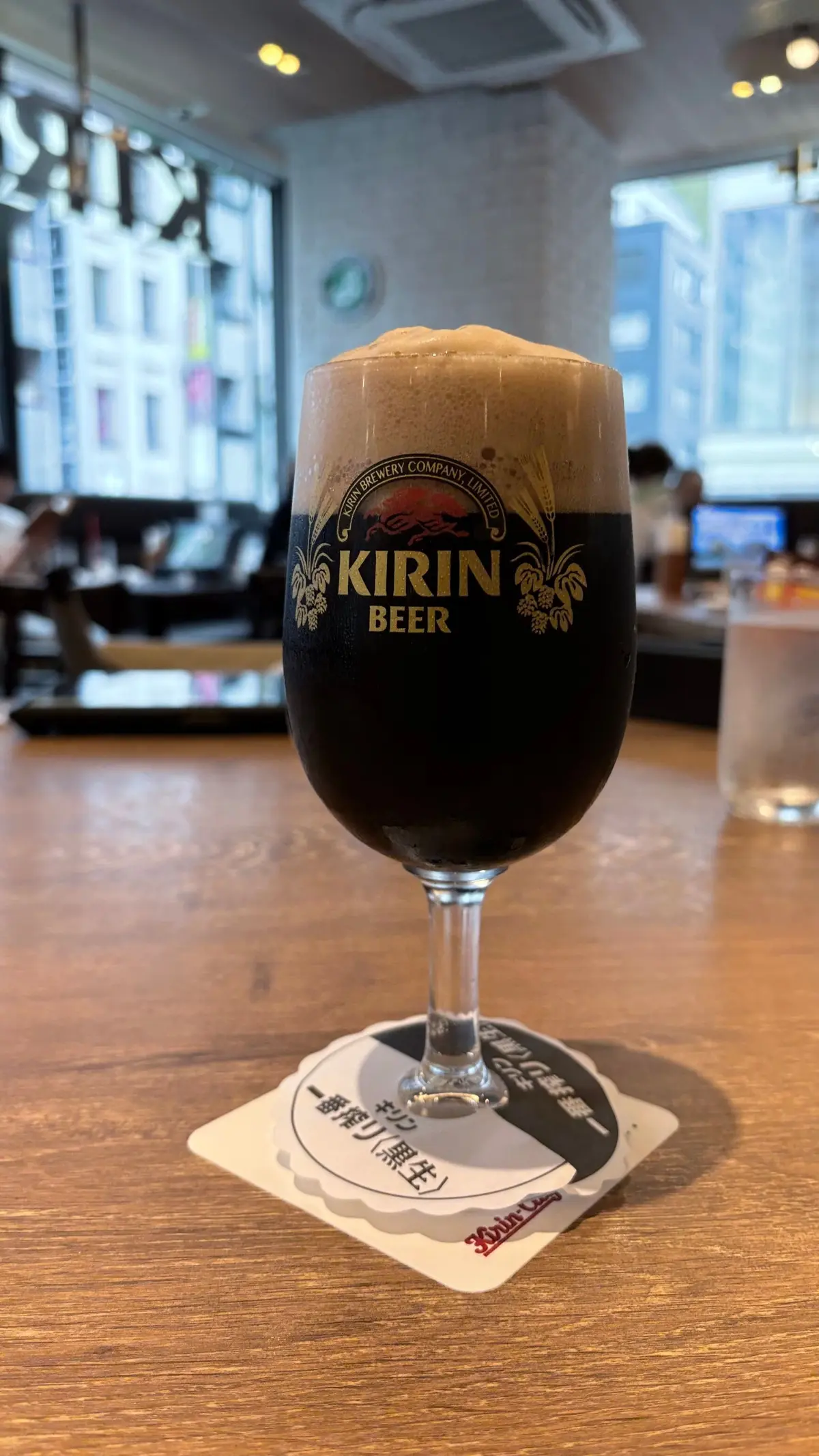 木製のテーブルに置かれた、キリンビールの黒ビール2杯。グラスにはきめ細かい泡が乗っており、背景には窓から見える景色と、レストランの様子が写っている。