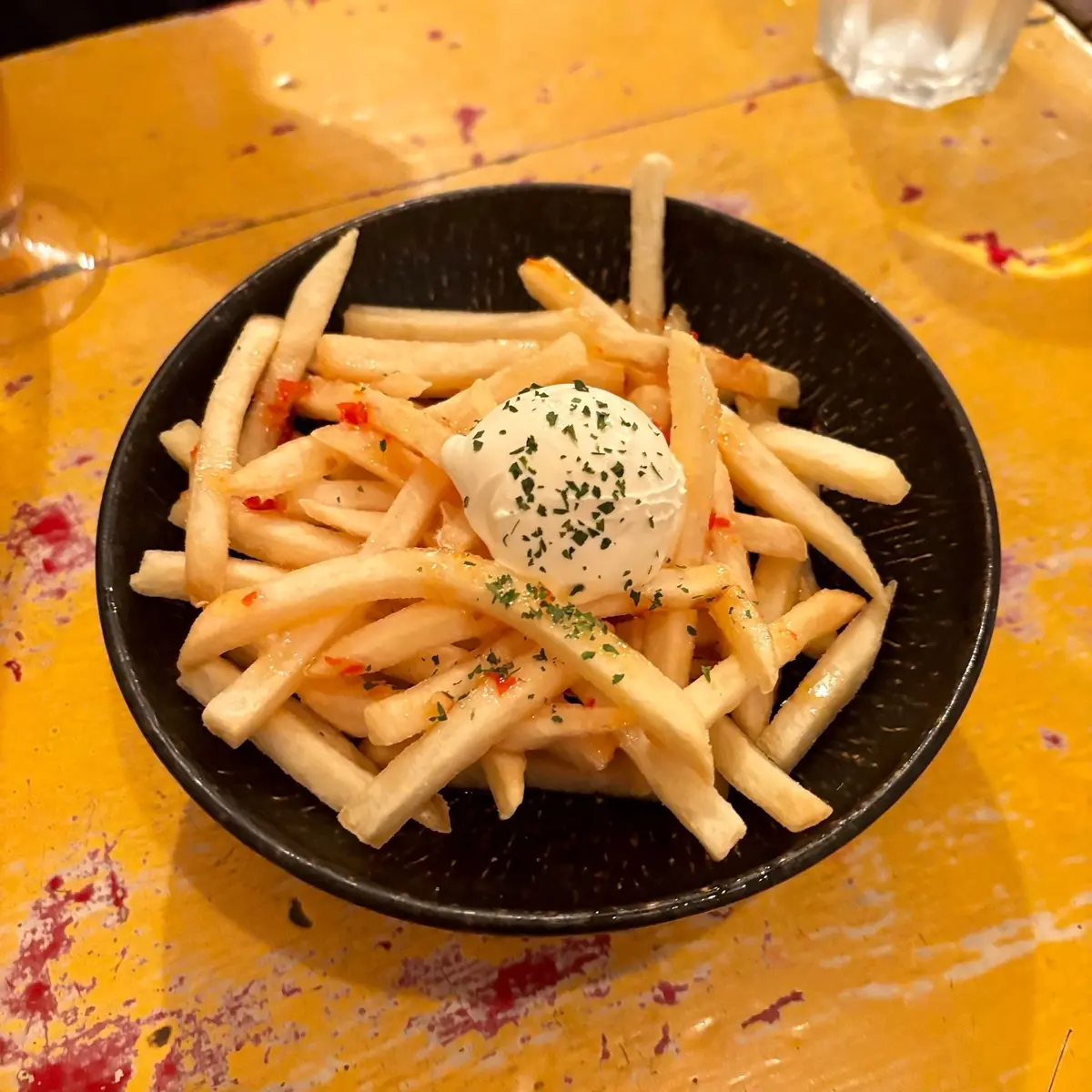 黒いボウルに入ったフライドポテト。ポテトの上にはクリームチーズが乗っており、赤唐辛子とパセリが散らされている。背景は黄色いテーブル。