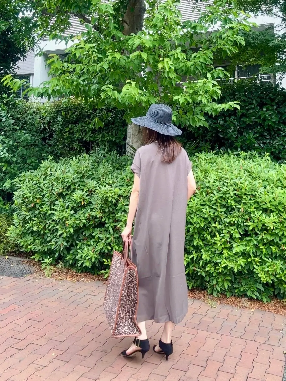 グレーのロングドレスと帽子を被った女性が、ブッシュの生い茂る庭を歩き、ヒョウ柄のトートバッグを手にしています。