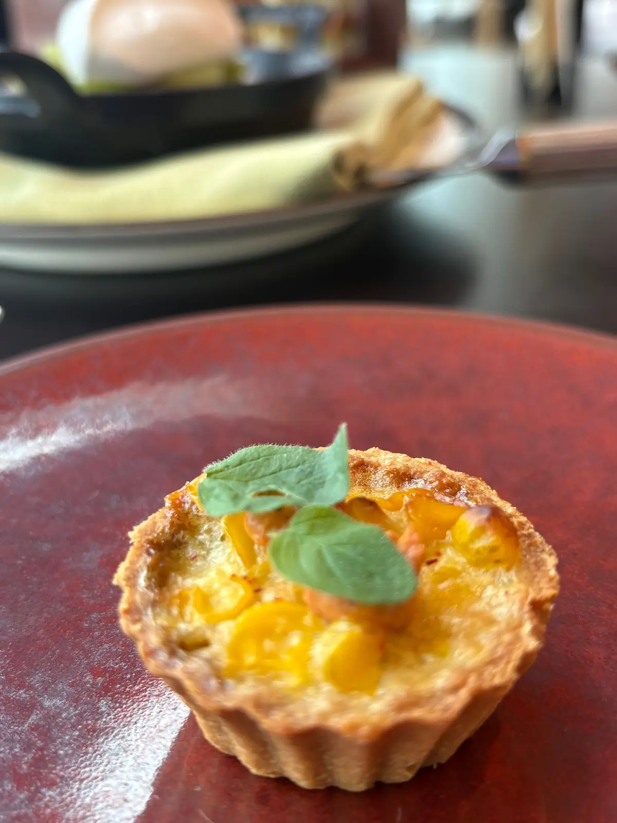 赤い皿に盛り付けられた、小さな卵焼きのタルト。タルト生地に、黄色い卵焼きと赤色の具材が詰まっており、上には緑色のハーブが添えられている。背景には、黒い鉄製のフライパンに入った料理が見える。