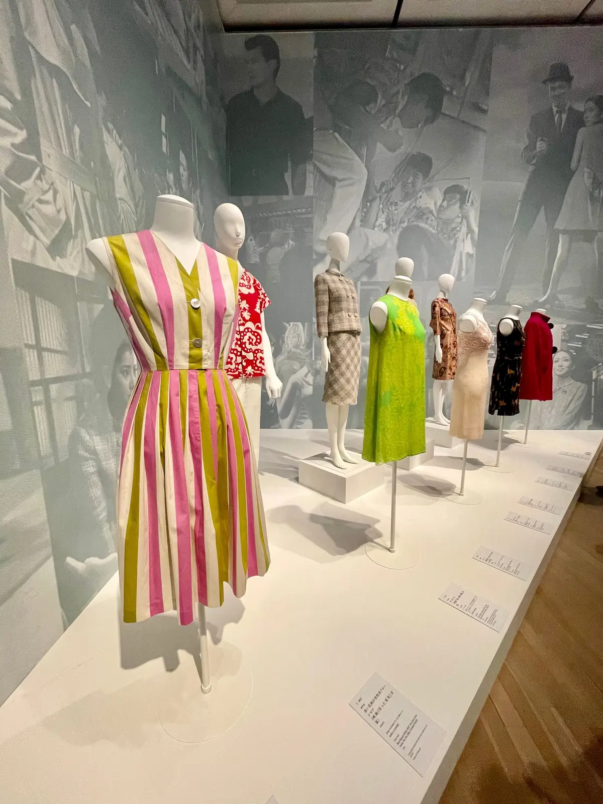 カラフルなストライプドレスや緑のワンピースなど、さまざまなデザインの衣服がマネキンに展示されたファッション展示。背景には歴史的な写真が飾られている。