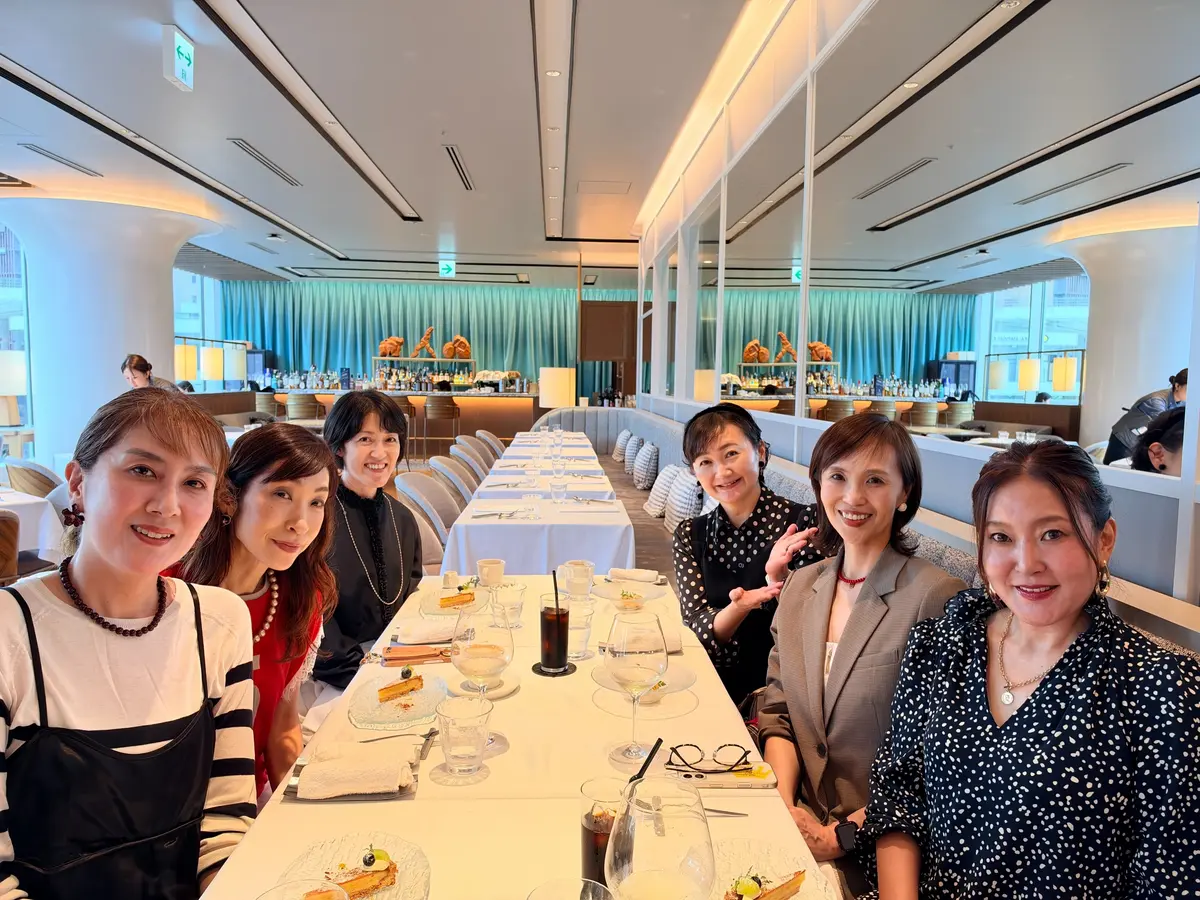 白を基調としたレストランで、7人の女性がテーブルを囲んで笑顔。テーブルにはグラスや料理が並び、背景に青色の装飾が見える。