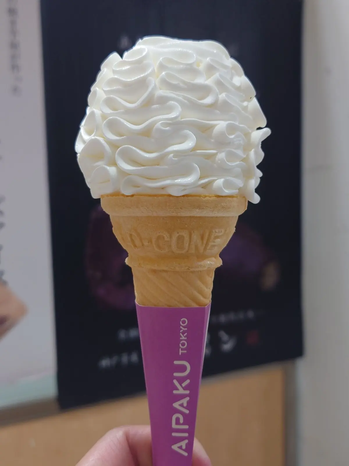 白いソフトクリームがワッフルコーンに乗ったアイスクリーム。ピンク色のスティックには「Aipaku Tokyo」のロゴがプリントされている。