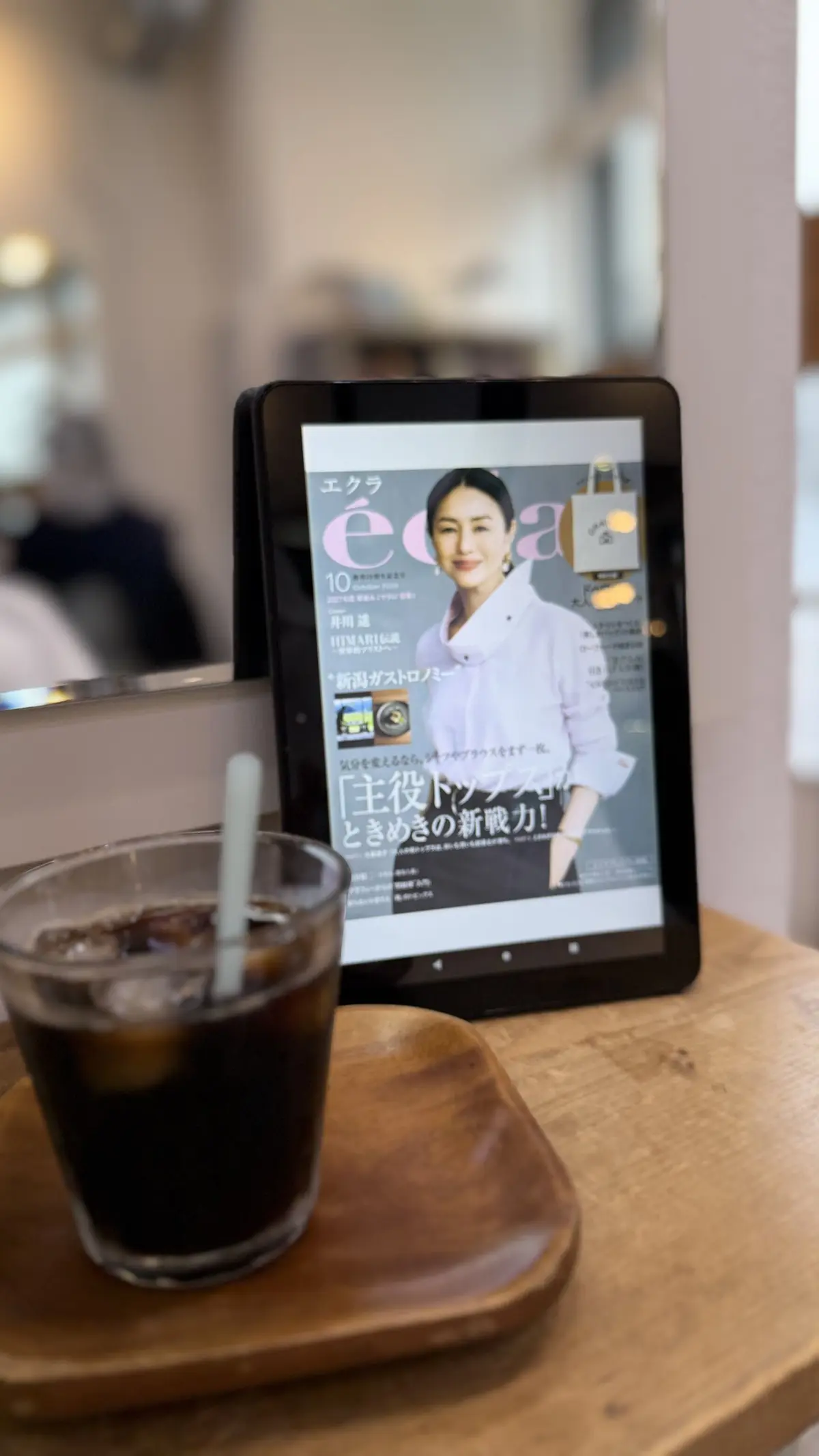 木製のテーブルに置かれたグラスの黒いコーヒーと、電子書籍リーダー。リーダーには日本の雑誌「エクラ」の表紙が表示されており、女性のポートレートが写っている。背景にはカフェの窓と席が見える。