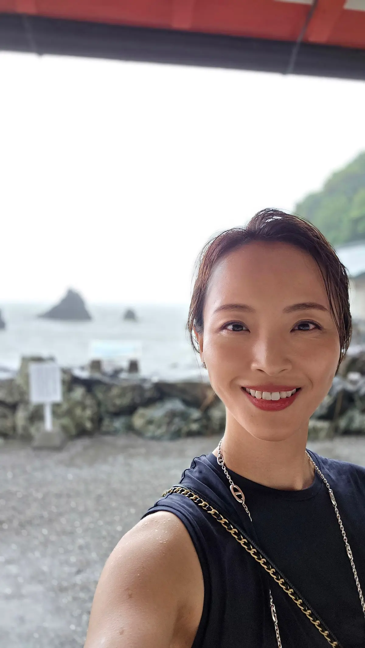 海を背景に、笑顔の女性が自撮りをしている写真。黒いトップスを着用し、髪を高い位置でまとめている。赤い屋根の建物が上部に写り、背景には岩場と海、遠くに島が見える。