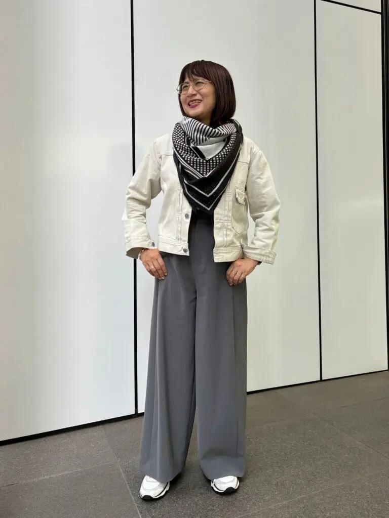 白いデニムジャケットに、黒と白の幾何学模様のスカーフを巻いた女性。グレーのワイドパンツと白いスニーカーを着用。笑顔でこちらを見ている。背景は白い壁。
