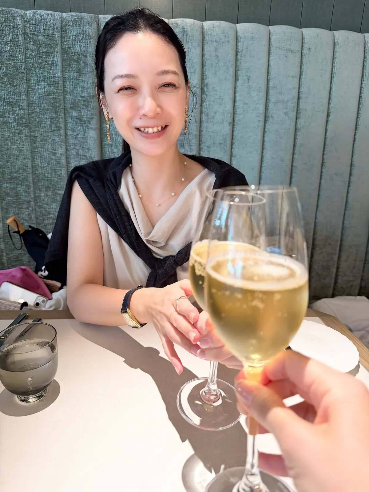 レストランのテーブルで、女性がグラスを手に笑顔を見せている。背景には青緑色の壁があり、テーブルには食器が置かれている。