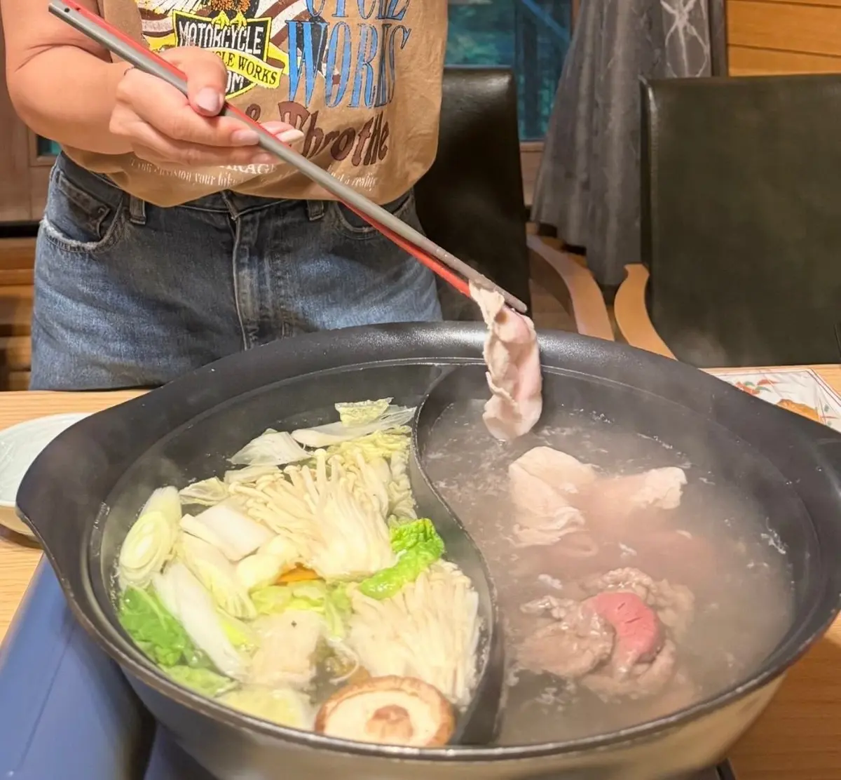 鉄製の鍋で煮込まれた豚肉や野菜が湯気を上げている様子。男性がトングで豚肉を持ち上げている。背景には椅子と窓が見える。