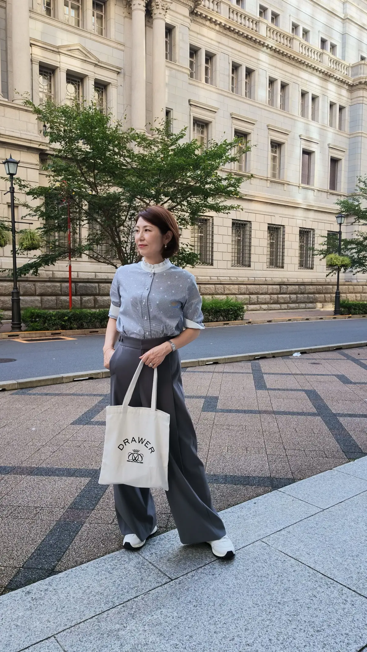 グレーのトップスとワイドパンツを着用した女性が、白いトートバッグを提げ、街並みを歩いている。背景にはクラシックな建物がある。