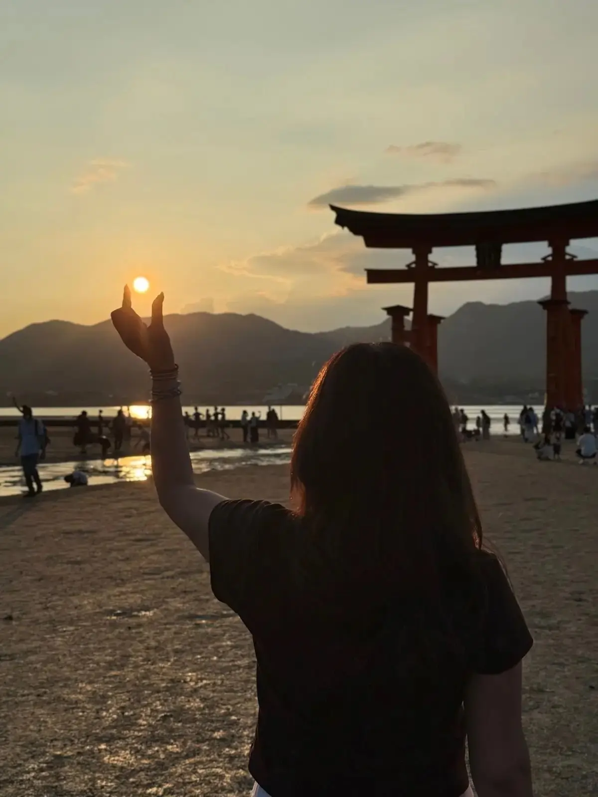 夕焼けの中、海辺に立つ人影。右手に太陽を掴むように手を伸ばし、背景には赤い鳥居がそびえ立つ。多くの人が水際で景色を眺めている。