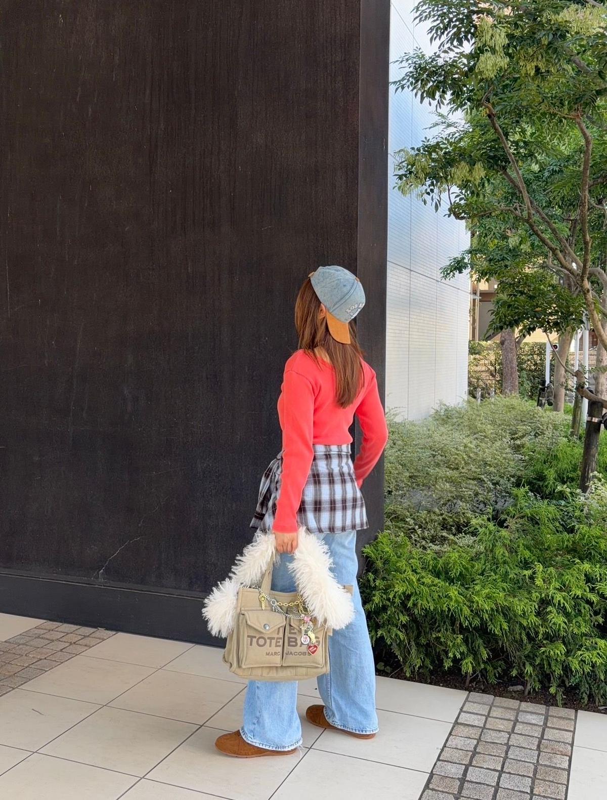 女性が後ろを向いて立っている写真。赤いニットにチェック柄のシャツ、ブルーのニット帽を着用。ベージュのトートバッグ（マーク・ジェイコブス）と茶色のブーツを持つ。背景には黒い壁と白い建物、緑の植栽がある。