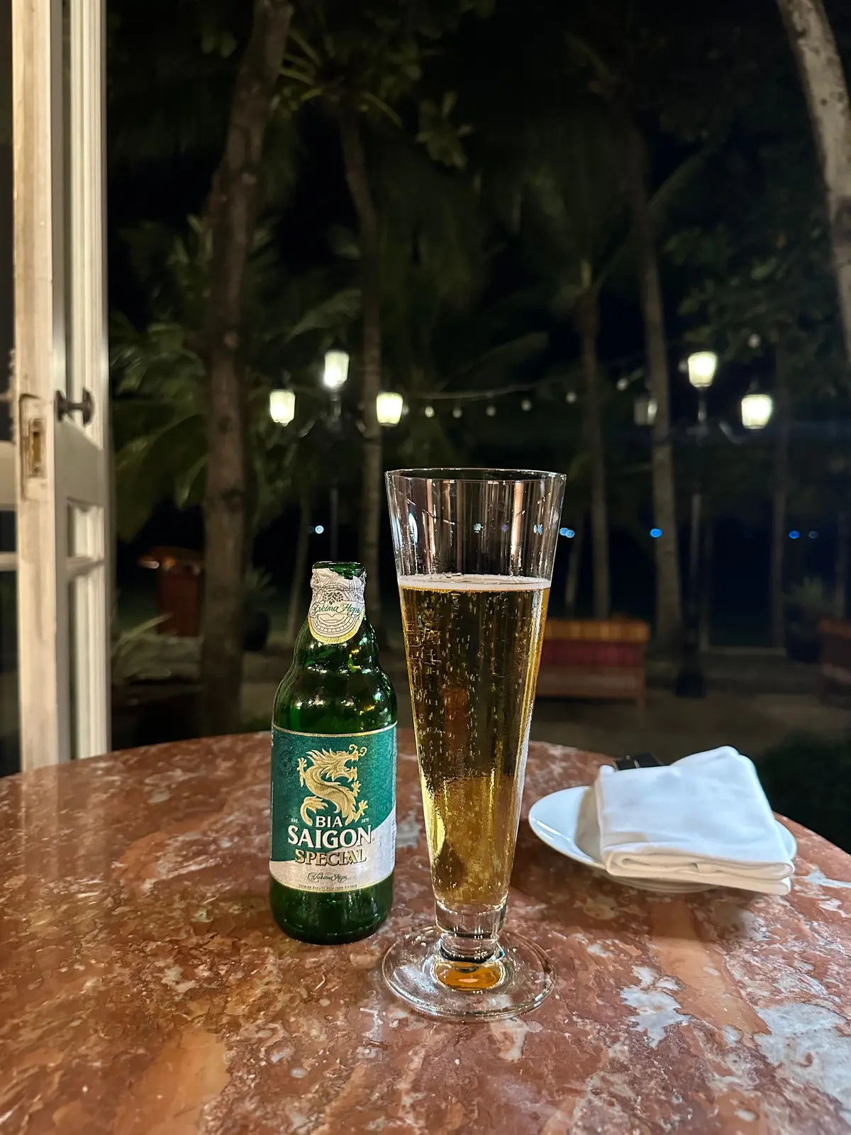 テーブルに置かれた、緑色の瓶の「サイゴンビール」とグラスに入ったビールのセット。背景には、ライトアップされた緑豊かな木々が見える。木々の後ろには漆黒の海が広がっている。