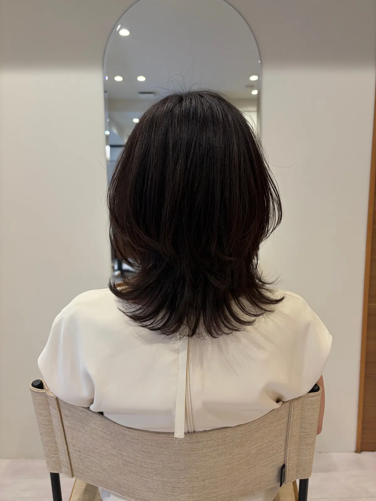 黒髪の女性が白いトップスを着て椅子に座っている後ろ姿。首の後ろから肩にかけて、レイヤーカットされた髪がふんわりと広がっている。背景には、丸い鏡とシンプルな内装が見える。