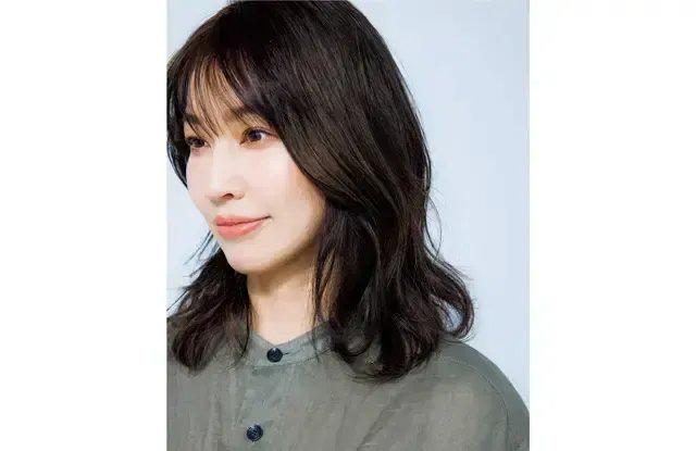 女性が淡い緑色の服を着て、ほほにほくろがある。黒髪は波打ったスタイルで、左肩にかけている。