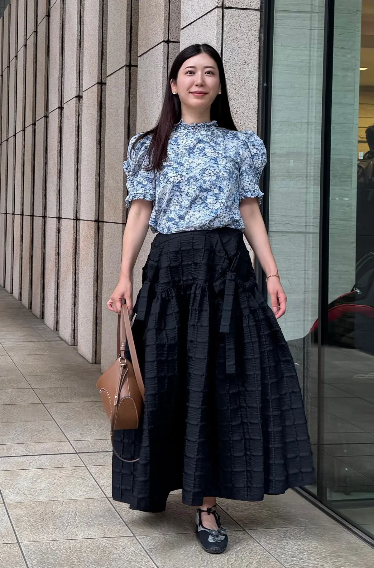 青と白のフラワープリントのプルオーバーと黒のフレアスカートを着た女性が、ガラスの建物前に立ち、茶色のハンドバッグを左手に持ち、笑顔でカメラを見つめている。