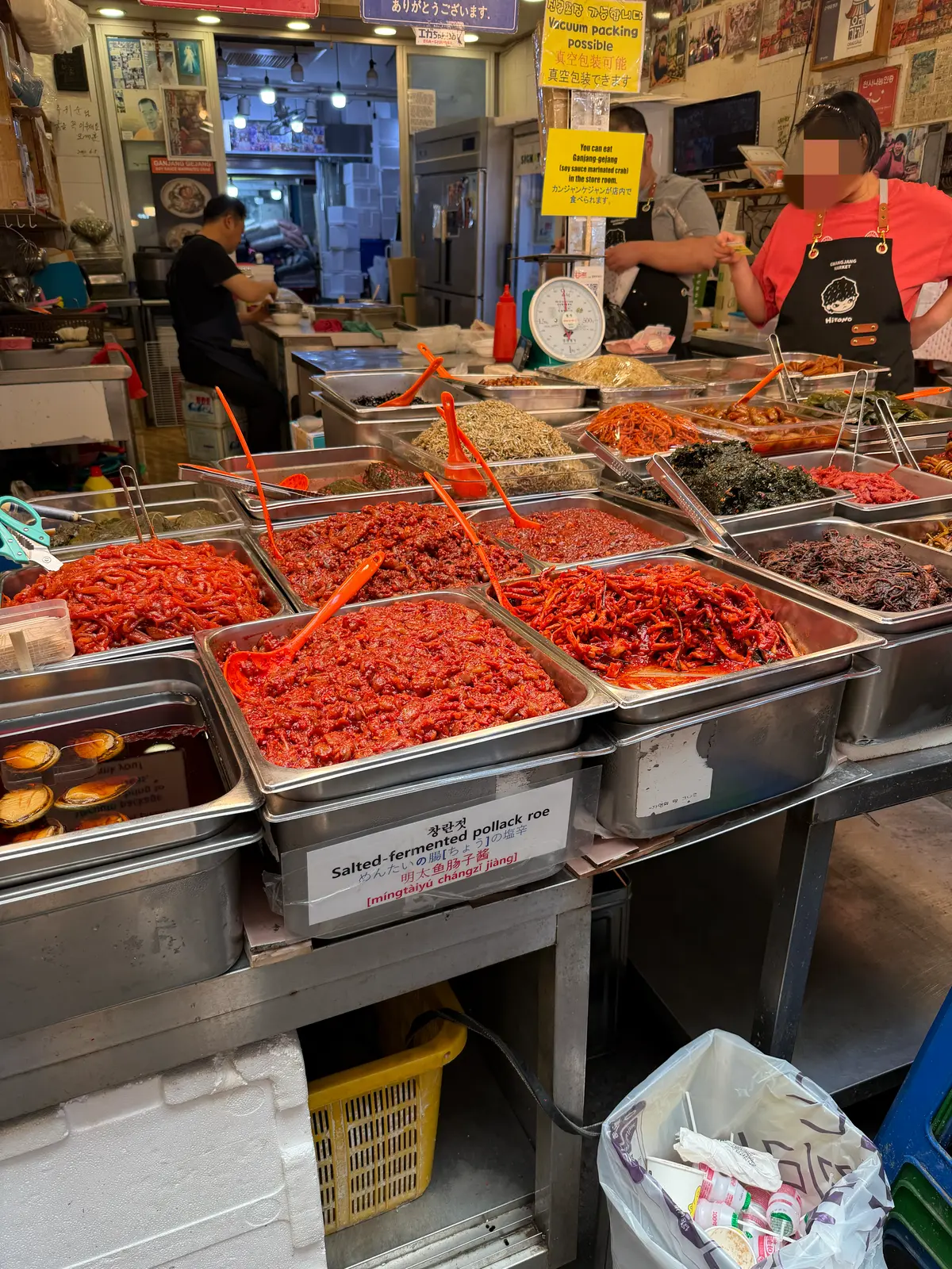 多数の金属トレイに赤い辛い野菜や海藻が並び、店員が作業する市場の屋内カウンター