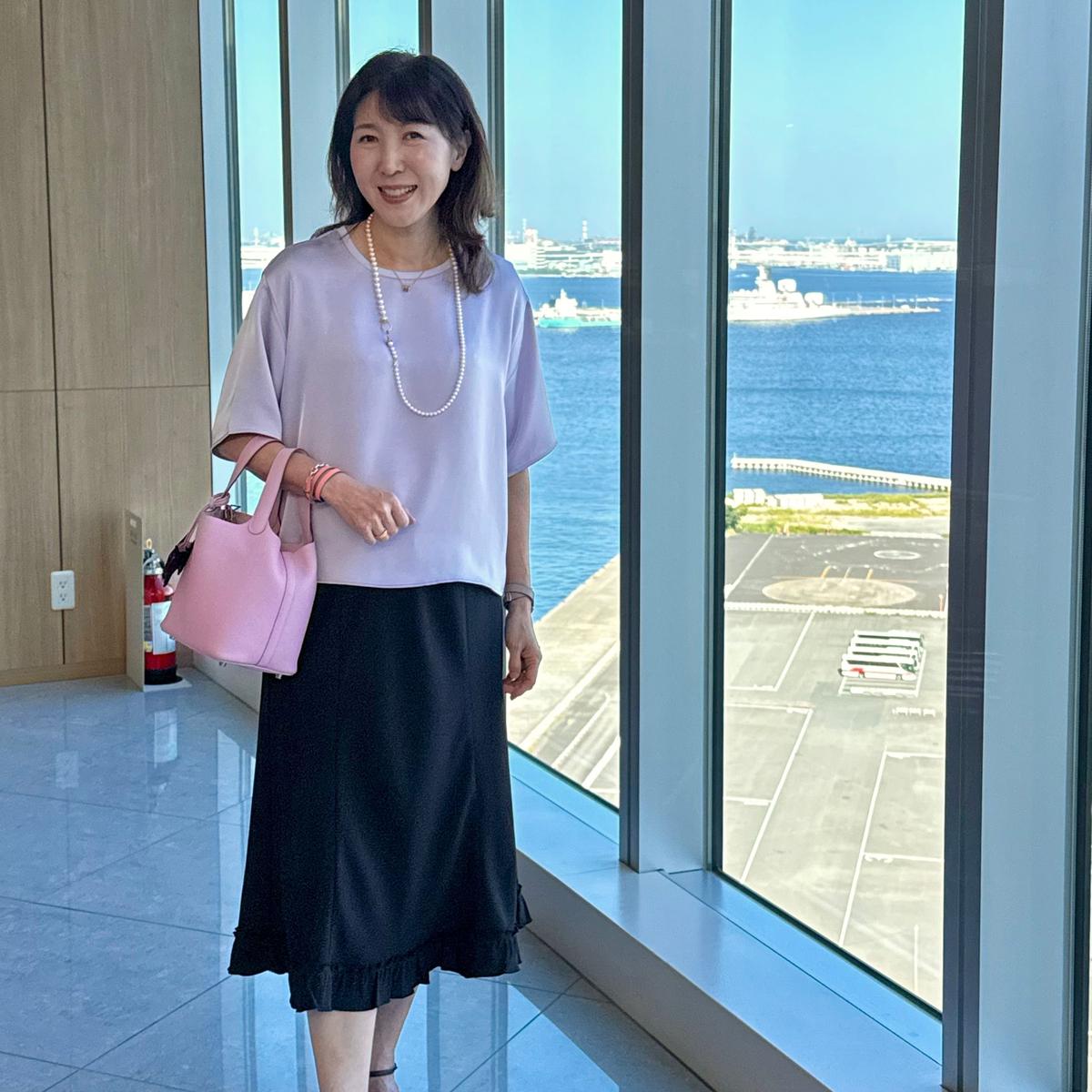 窓際に立つ女性。淡い紫色のトップスに黒いフレアスカート、パールネックレスを着用。右手にピンク色のハンドバッグを持ち、微笑んでいる。背景には青い海と港の風景が広がる。