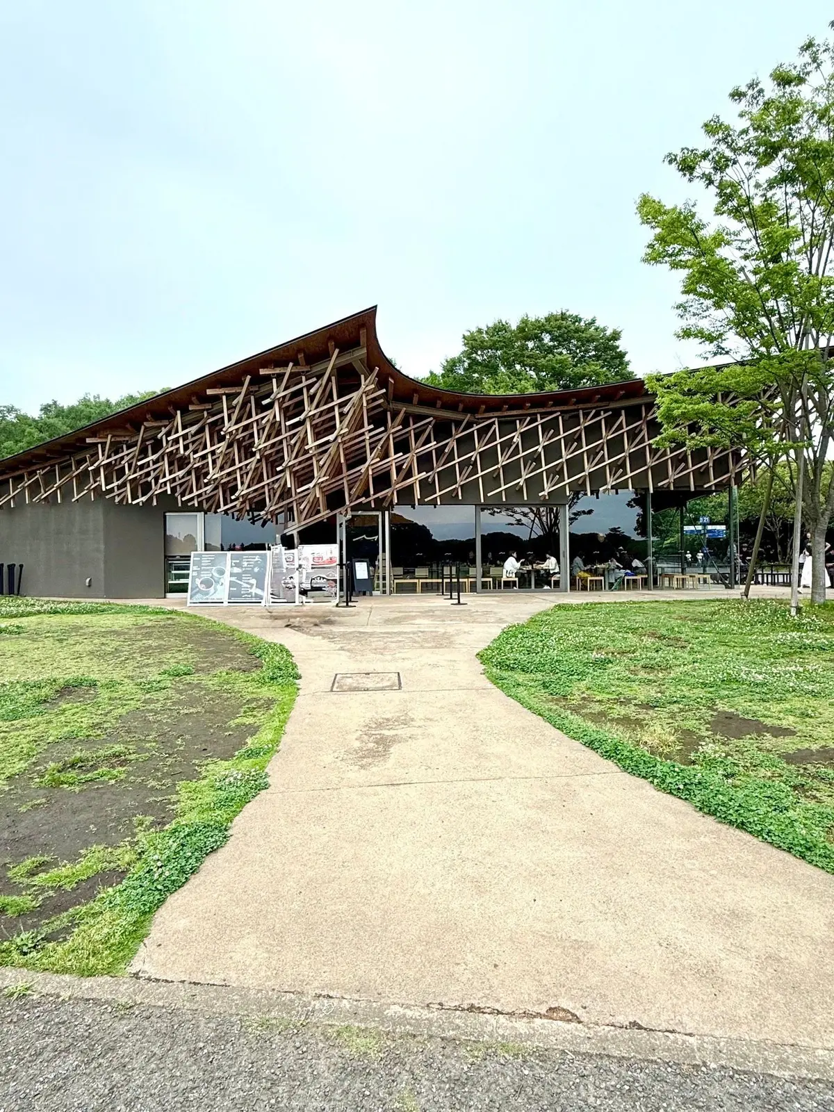 国営昭和記念公園、みんなの原っぱ、オカカフェ、木製の屋根とコンクリート壁のモダンなカフェ、芝生と木々のある屋外空間、テーブルと椅子に座る人々
