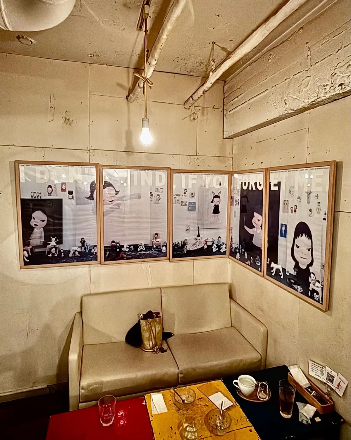 奈良美智さんの絵画が5枚、壁に沿って配置されたコーナー席の背景に飾られている。席にはベージュのソファと、木製のテーブルが置かれ、飲み物や食器が置かれている。天井には配管が露出しており、電球が吊り下げられている。