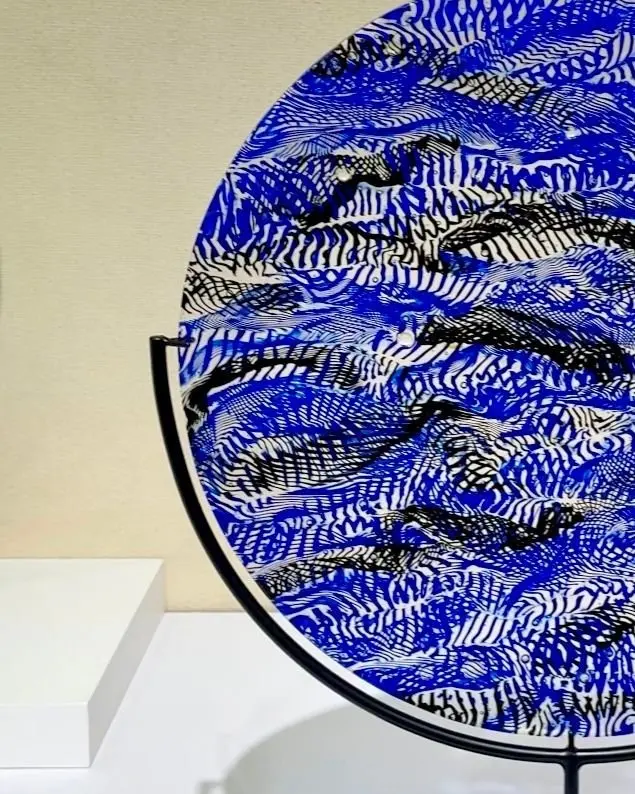 青地に白の模様が描かれた皿が、黒いスタンドに飾られている。皿は円形で、複雑な線描の模様が全体に広がっている。背景はベージュ色の壁。落書きシリーズ