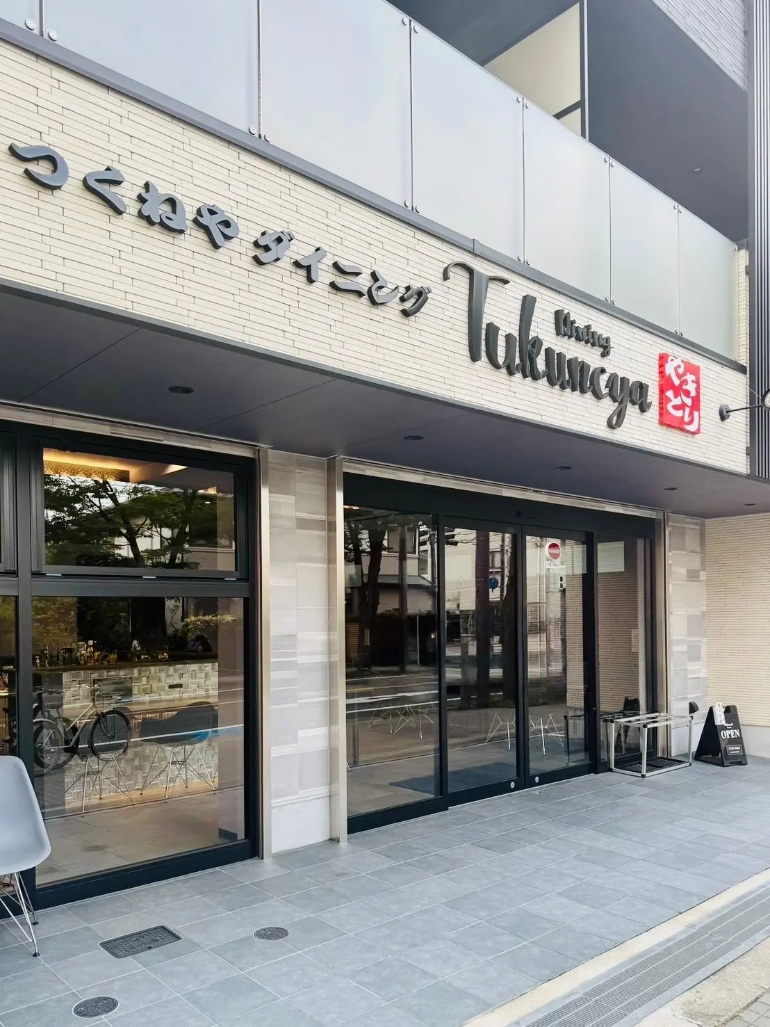 タイル張りの壁に日本語の看板付きの店入り口、ガラスドアと窓から店内が見え、椅子と自転車が映る