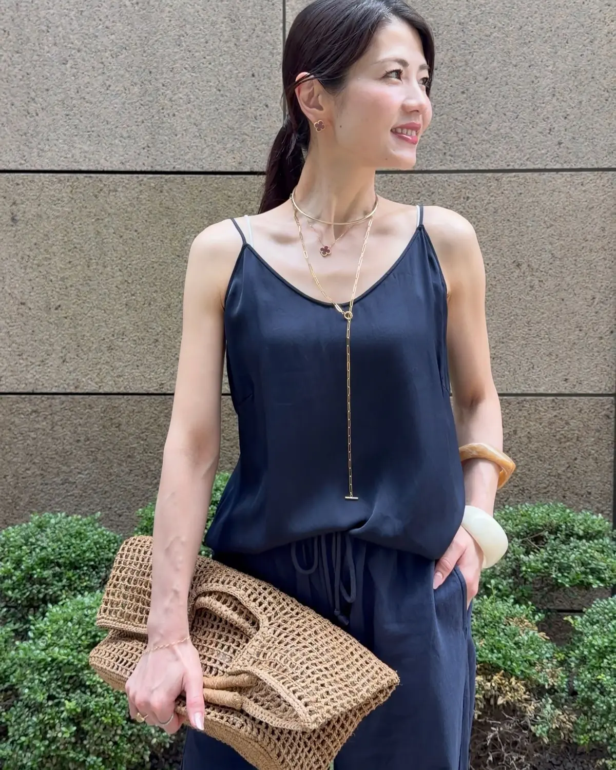 黒いキャミソールワンピースを着た女性。茶色の編み込みバッグを持ち、ゴールドのネックレスとイヤリングを身に着けている。背景にはコンクリートの壁と緑の植栽がある。