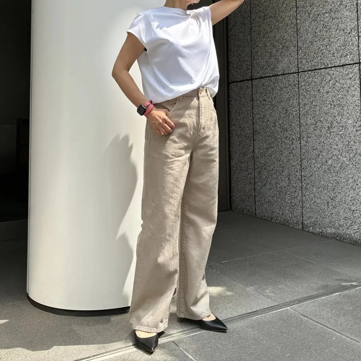 白いTシャツにベージュのワイドパンツを着用した女性が立っている全身写真。黒いフラットシューズを履き、左腕に腕時計をしている。背景には白い円柱とグレーの壁がある。